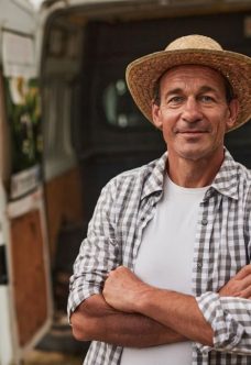 Smiling mature male farmer in straw hat standing with crossed arms against blurred van prepared for delivery of products at local market